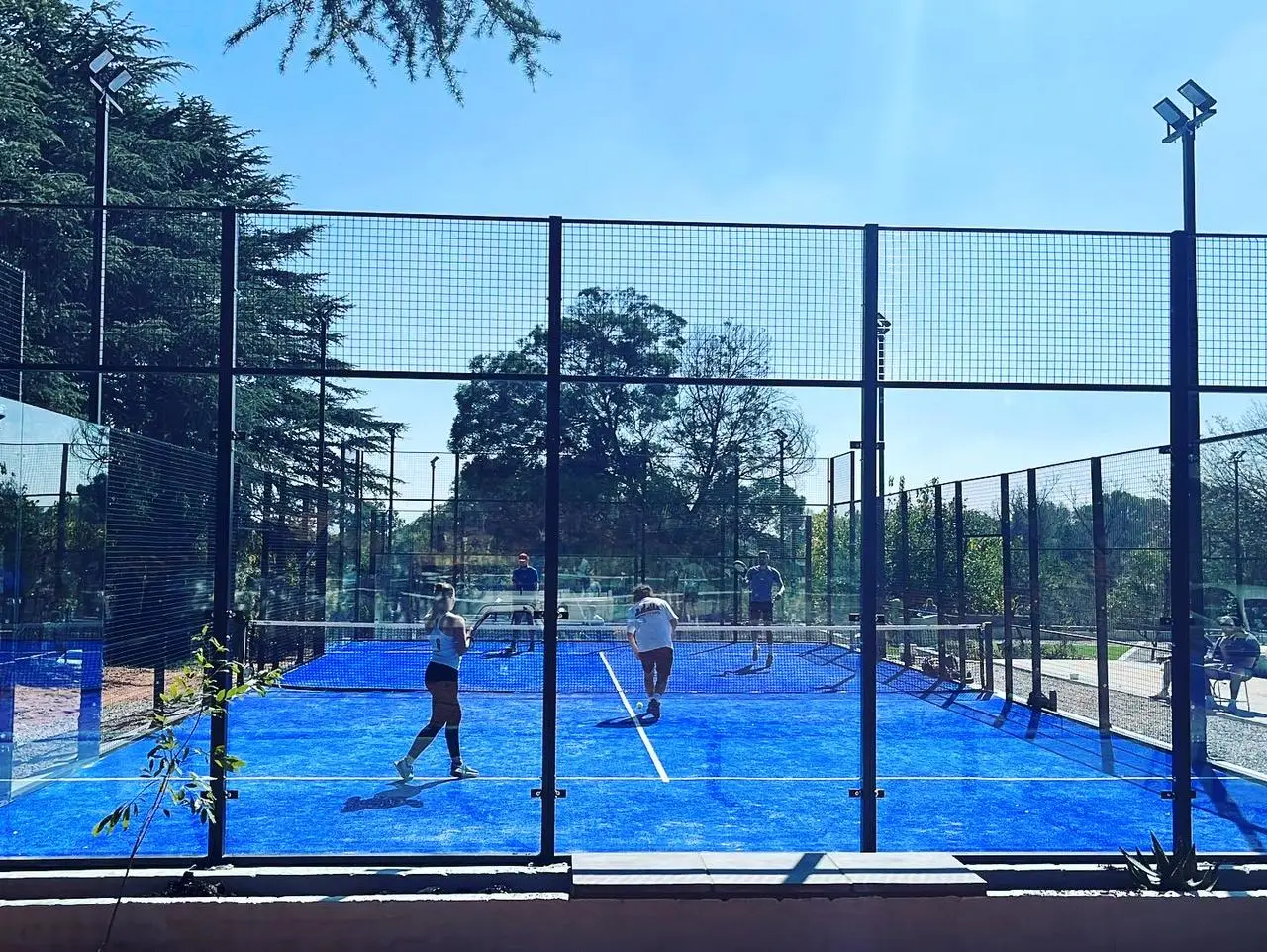 Two players engaged in a padel match on a blue court enclosed by a fence with trees in the background.