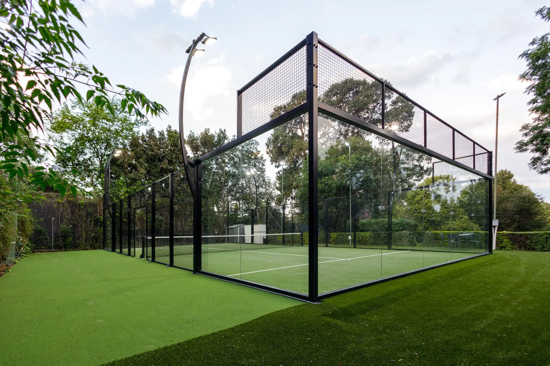 A modern paddle tennis court featuring glass walls and artificial turf, surrounded by greenery.