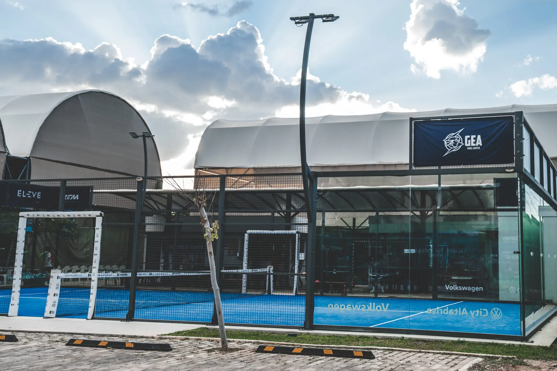 Exterior view of a glass paddle tennis court with a logo on the wall and blue court surface.