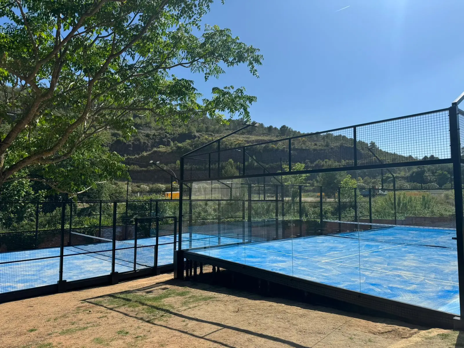 Two outdoor padel courts with blue flooring and black fencing in a natural setting.