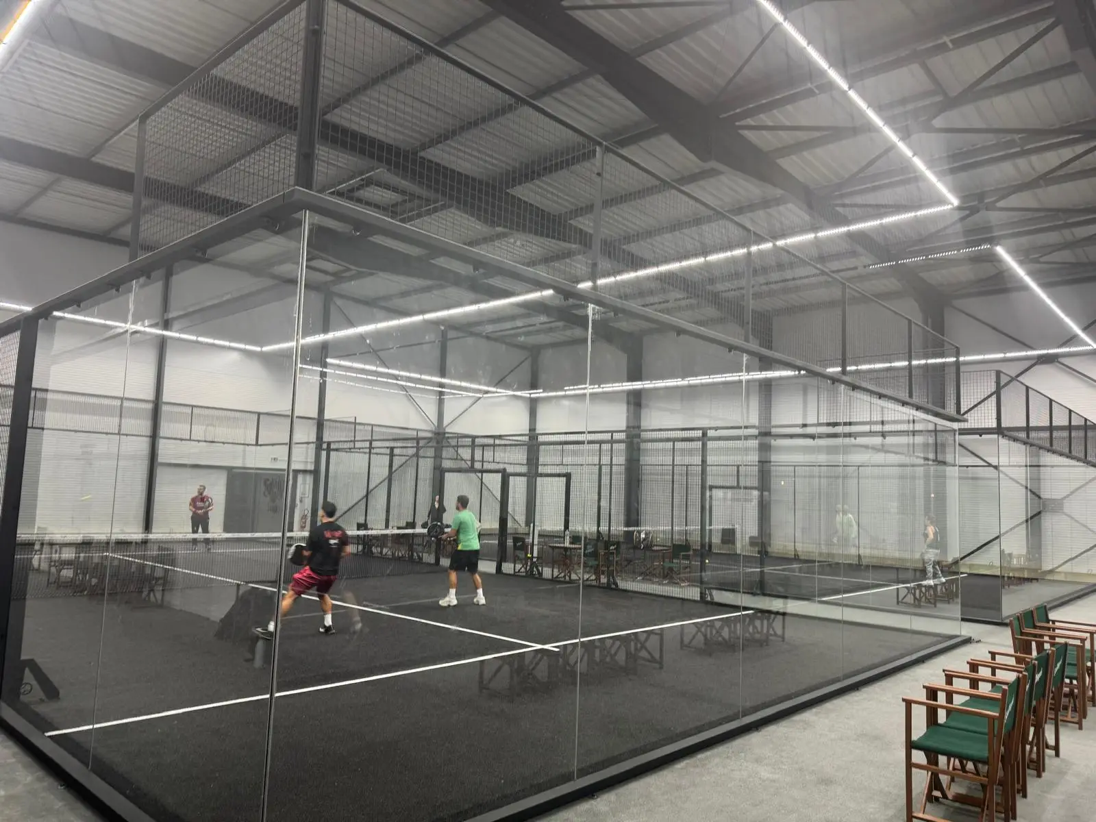 Two paddle tennis players competing in a glass-enclosed indoor court with additional courts visible.