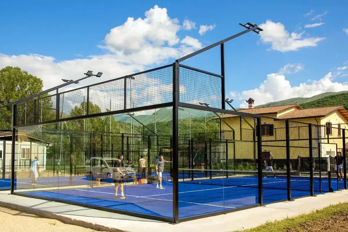 Players rally on a blue outdoor panoramic padel court with glass walls and black steel framing beside a school-style building under a bright sky.