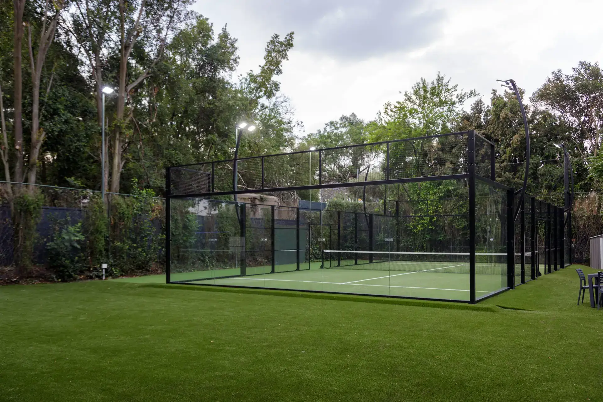 A standard outdoor padel court with glass walls and LED lighting sits beside lush trees, reflecting the budget-friendly school facility setting in South Africa.