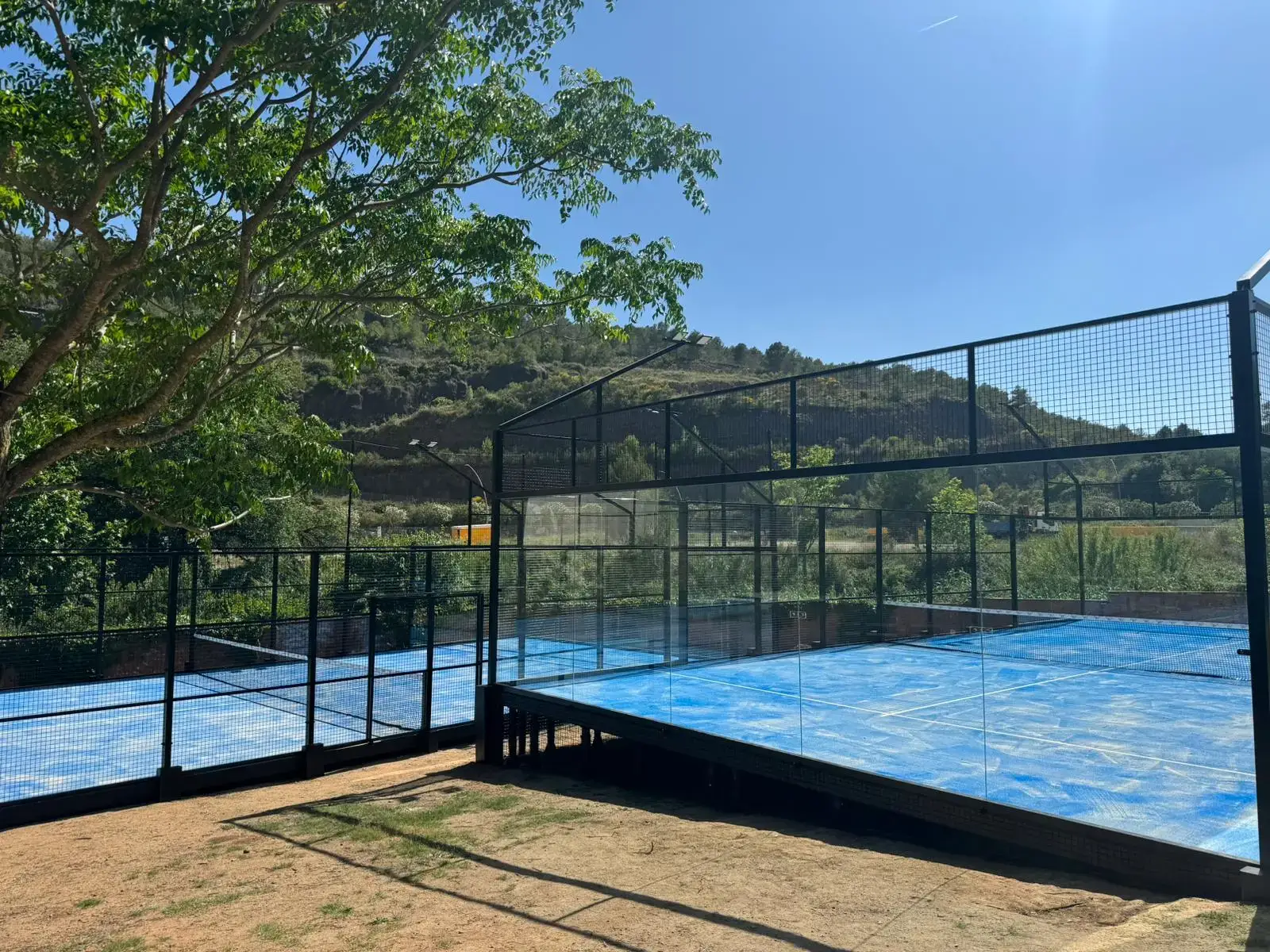 Two outdoor padel courts with blue flooring and black fencing in a natural setting